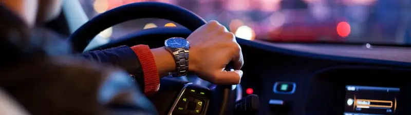 A driver's hands on the wheel at dusk, city lights ahead