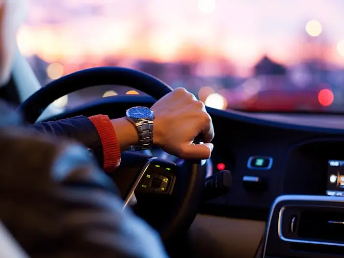 A driver's hands on the wheel at dusk, city lights ahead